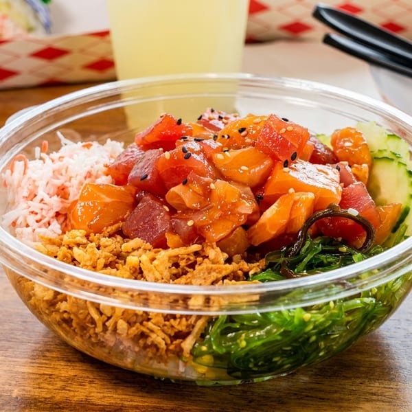 Vibrant poke bowl featuring diced fish, crab salad, seaweed, and crispy onions in a clear plastic bowl.