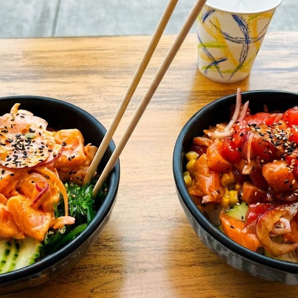 Two colorful poke bowls with fresh fish, corn, and vegetables, served with chopsticks on a wooden table.